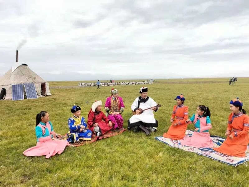 毡房外的草地上，孩子们围坐听长者演唱